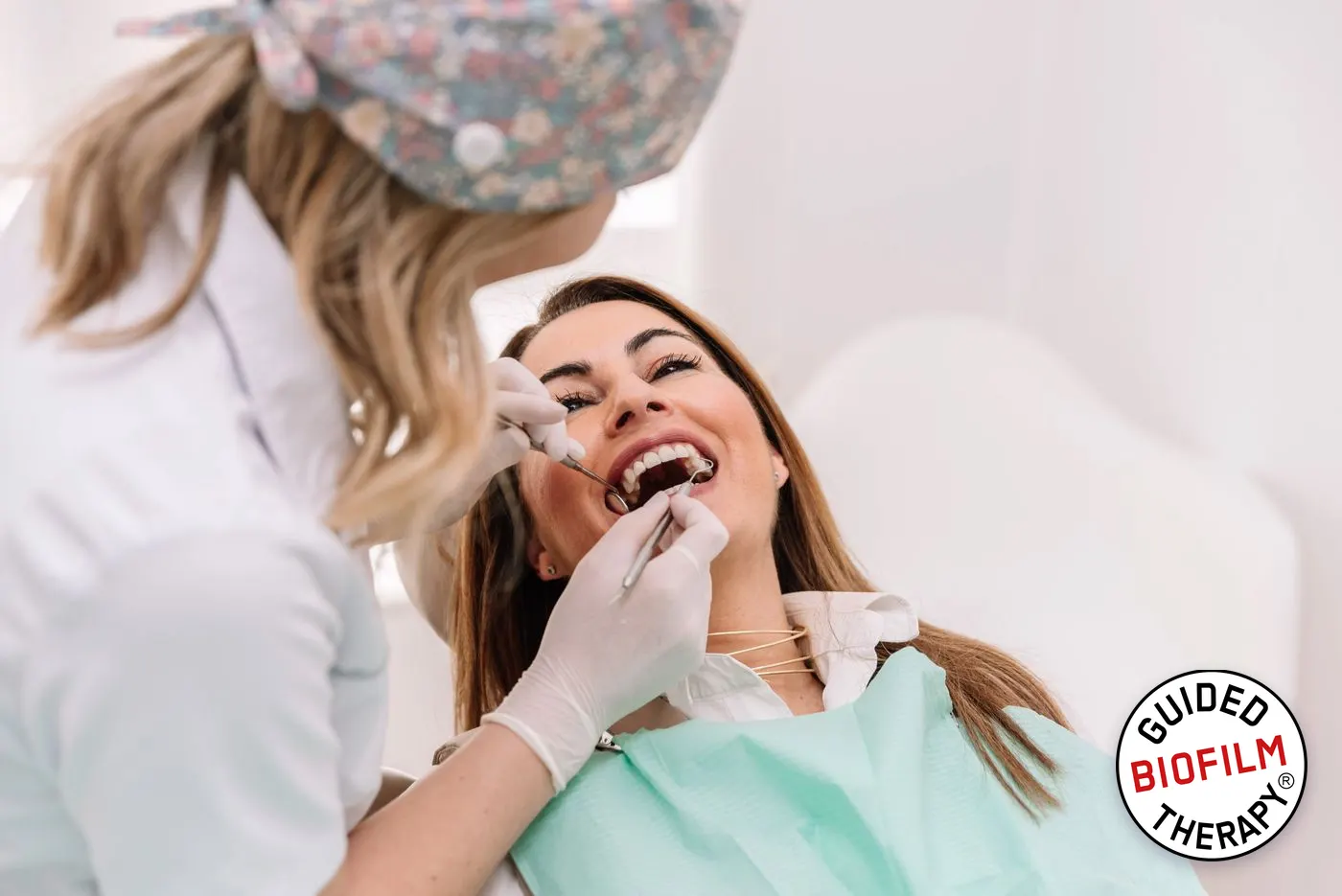 Mosolygó páciens rendszeres kontrollvizsgálaton, a fogorvos tükörrel és szondával ellenőrzi a fogakat; a sarokban a Guided Biofilm Therapy jelvény